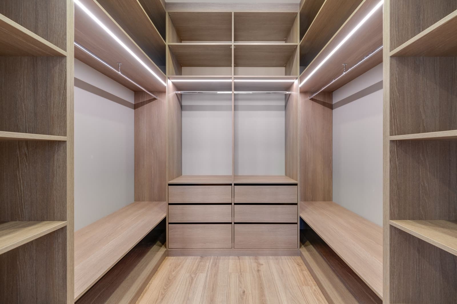 A walk-in wardrobe in light oak with open shelving, hanging rails, LED strip lighting and a central drawer unit.