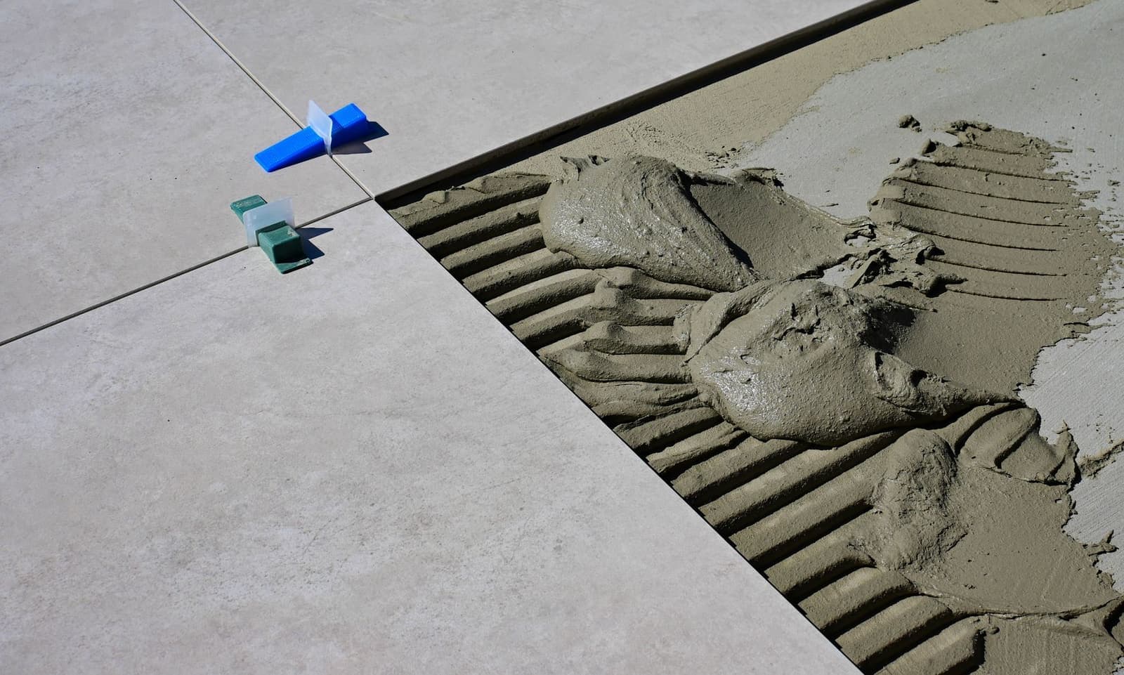 Large-format grey floor tiles being laid over combed adhesive, with tile levelling clips at the joints.