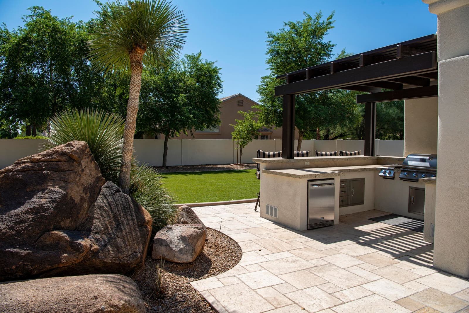 A paved patio with an outdoor kitchen under a pergola, beside a lawn, palm trees and a rock garden.