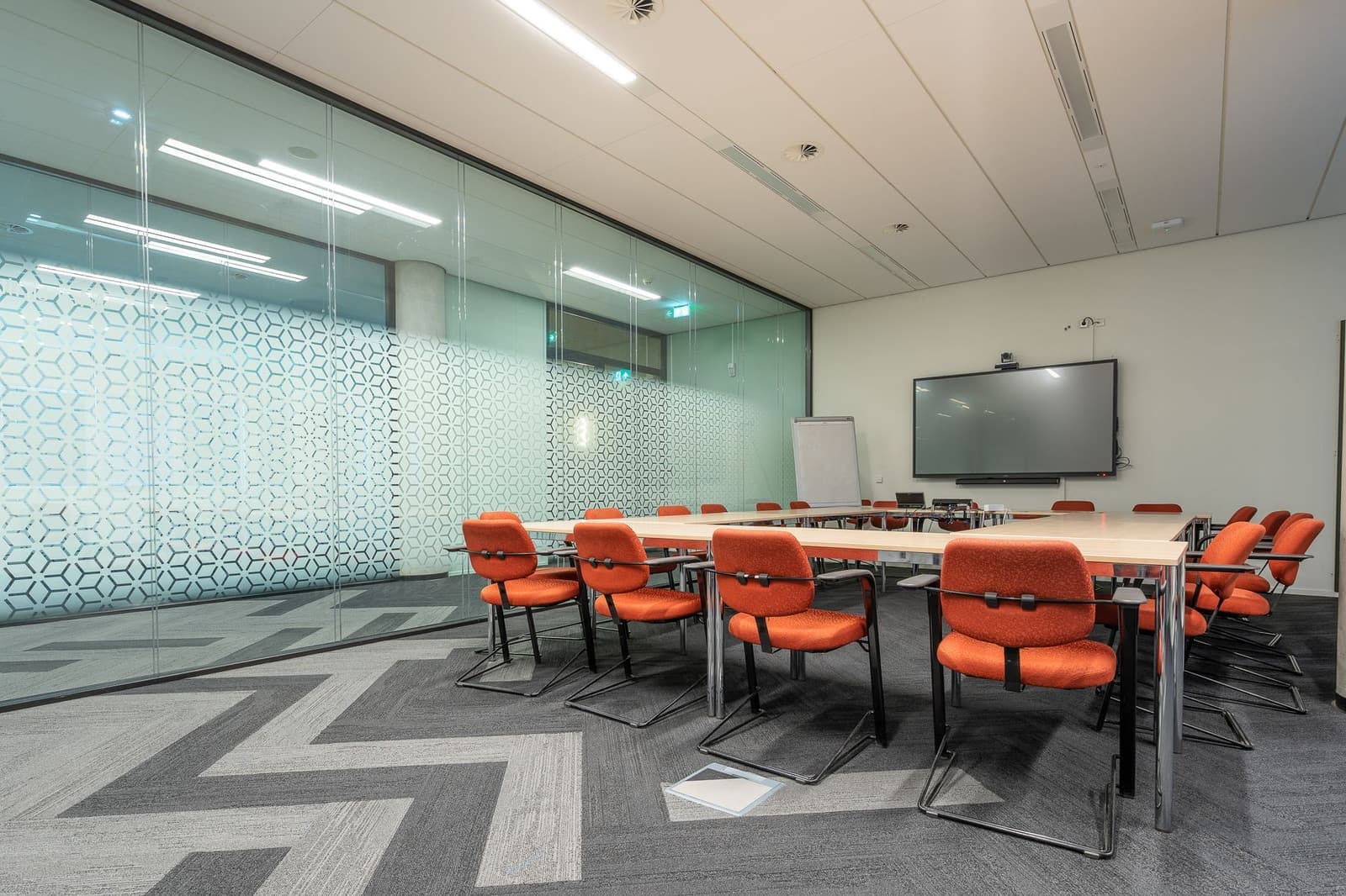 A meeting room enclosed by glass partitions with frosted manifestation, a long table, orange chairs and a wall screen.
