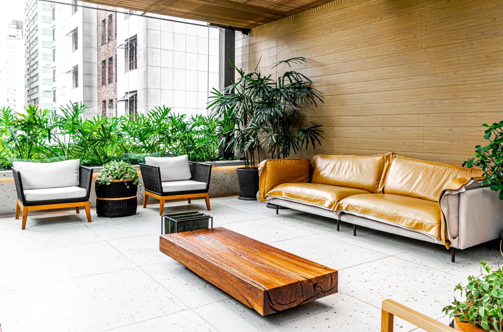 A covered terrace with a leather sofa, armchairs, a timber coffee table and planting beside high-rise buildings.