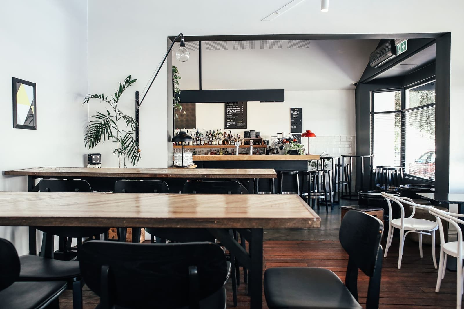 An empty cafe with timber tables, black chairs, a bar counter and plants by a large window.